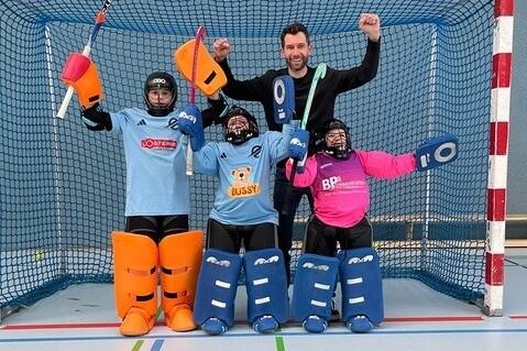 Gruppe von vier Personen in Sportausr&uuml;stung, die in einem Hockeytor posieren, ein Trainer hebt jubelnd die Arme.