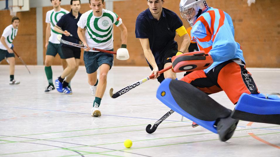 Mehrere Spieler in einem Hallenhockeyspiel, ein Torwart weicht einem Schuss in die Ecke aus. Gelbe Hockeykugel sichtbar.