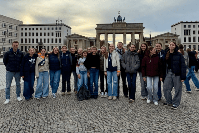 Gruppe von jungen Erwachsenen posiert l&auml;chelnd vor dem Brandenburger Tor, umgeben von historischen Geb&auml;uden.