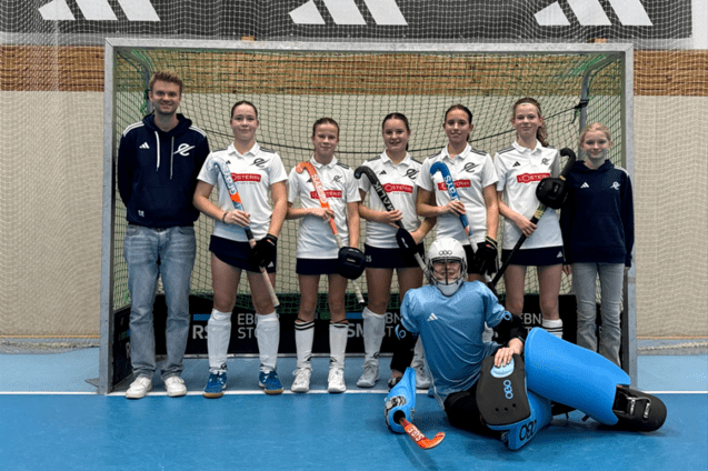 Teamaufstellung von sechs Hockeyspielerinnen und einem Trainer vor einem Tor, eine Spielerin in Torwartausr&uuml;stung sitzt vorne.