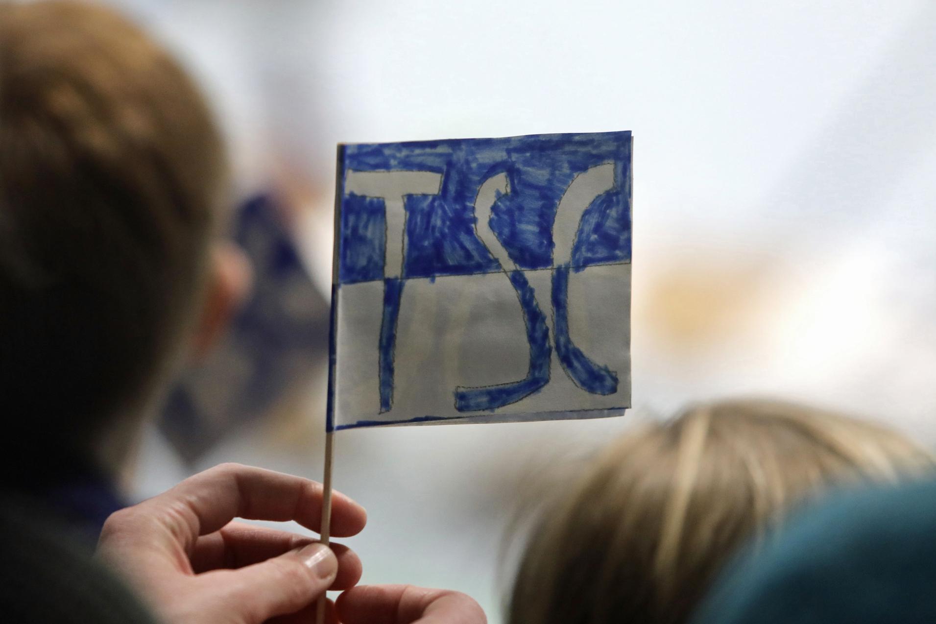 Eine Person h&auml;lt eine kleine, handgemalte Fahne mit der Aufschrift &bdquo;TSC&ldquo; in blauer Schrift vor einer Zuschauergruppe.