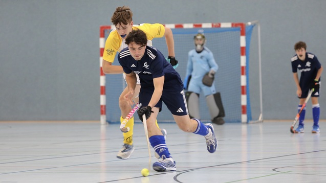 Zwei Hockeyspieler k&auml;mpfen um den Ball in einer Halle, im Hintergrund steht ein Torwart.