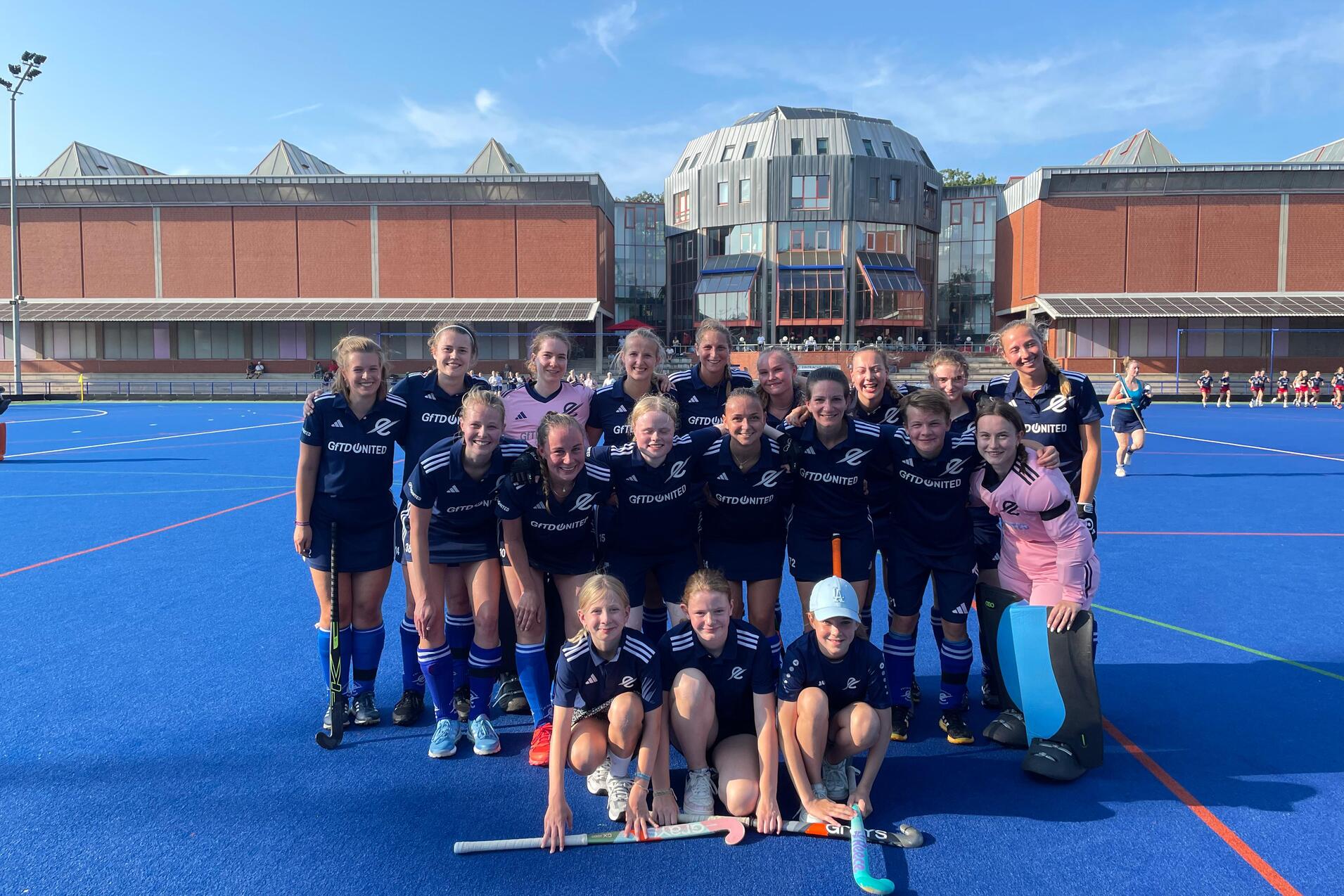 Gruppenfoto eines Hockeyteams in blauen Trikots auf einem Kunstrasenplatz, im Hintergrund moderne Geb&auml;ude und klare Himmel.
