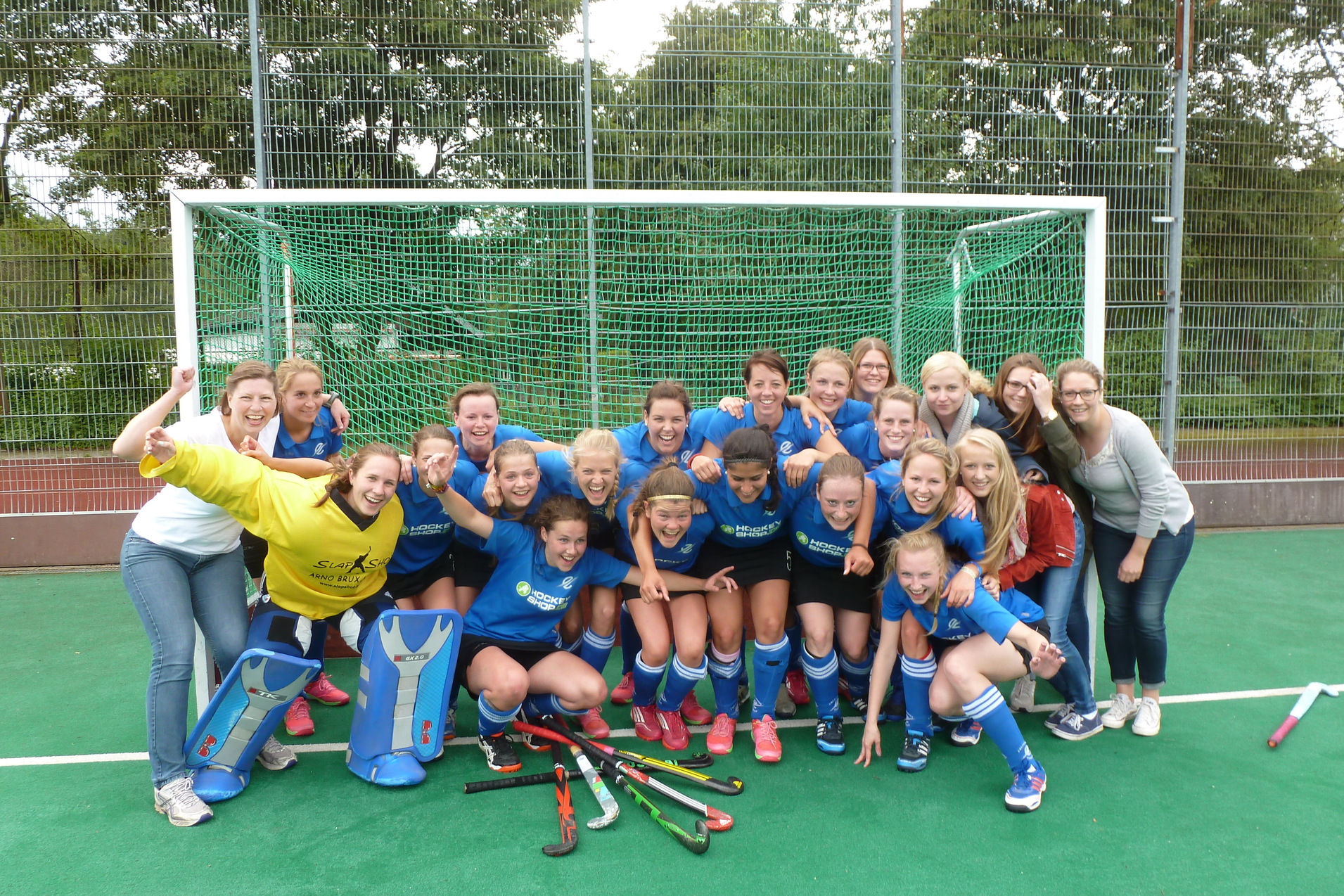 Gruppfoto einer Hockeymannschaft mit Trainerin, alle tragen blaue Trikots und stehen vor einem Hockeytor auf einem Kunstrasenfeld.
