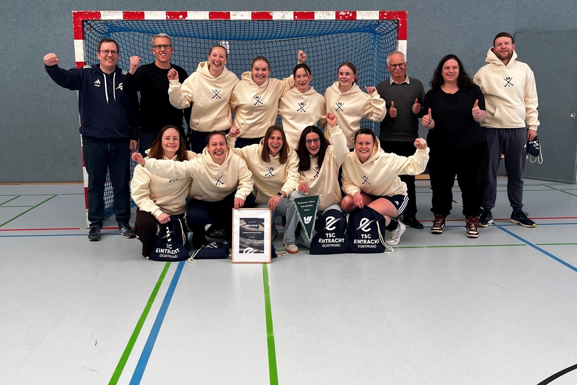 Gruppe von Handballspielern in wei&szlig;en Sweatshirts posiert vor einem Tor, l&auml;chelnd mit Pokalen und Taschen.