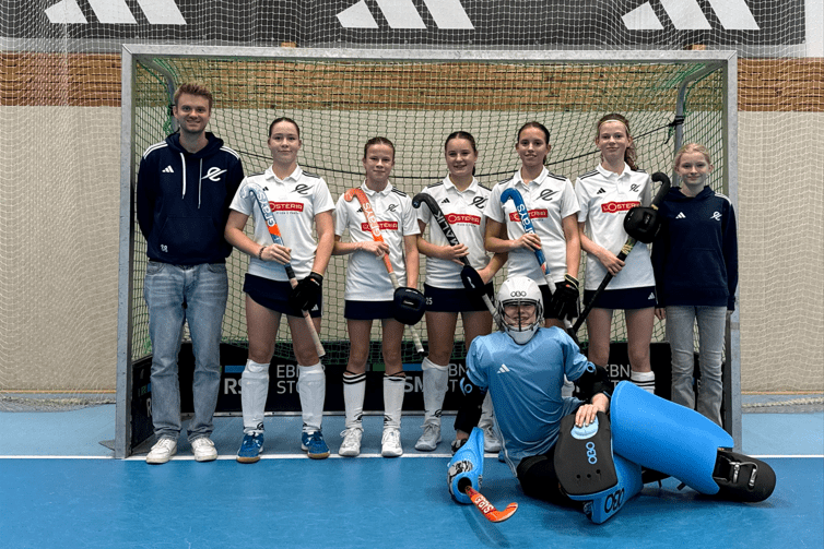 Teamaufstellung von sechs Hockeyspielerinnen und einem Trainer vor einem Tor, eine Spielerin in Torwartausrüstung sitzt vorne.