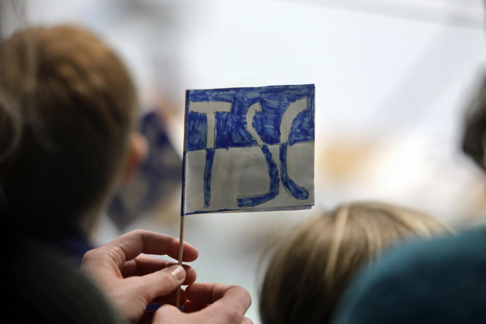 Eine Person hält eine kleine, handgemalte Fahne mit der Aufschrift „TSC“ in blauer Schrift vor einer Zuschauergruppe.