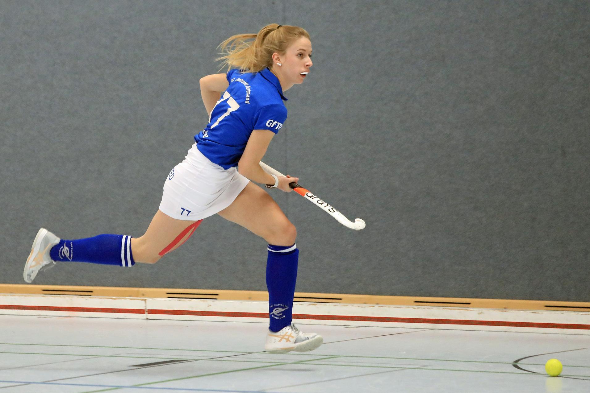 Spielerin in blauer Sportbekleidung sprintet mit dem Hockeyschläger in der Hand auf einen kleinen Ball zu.