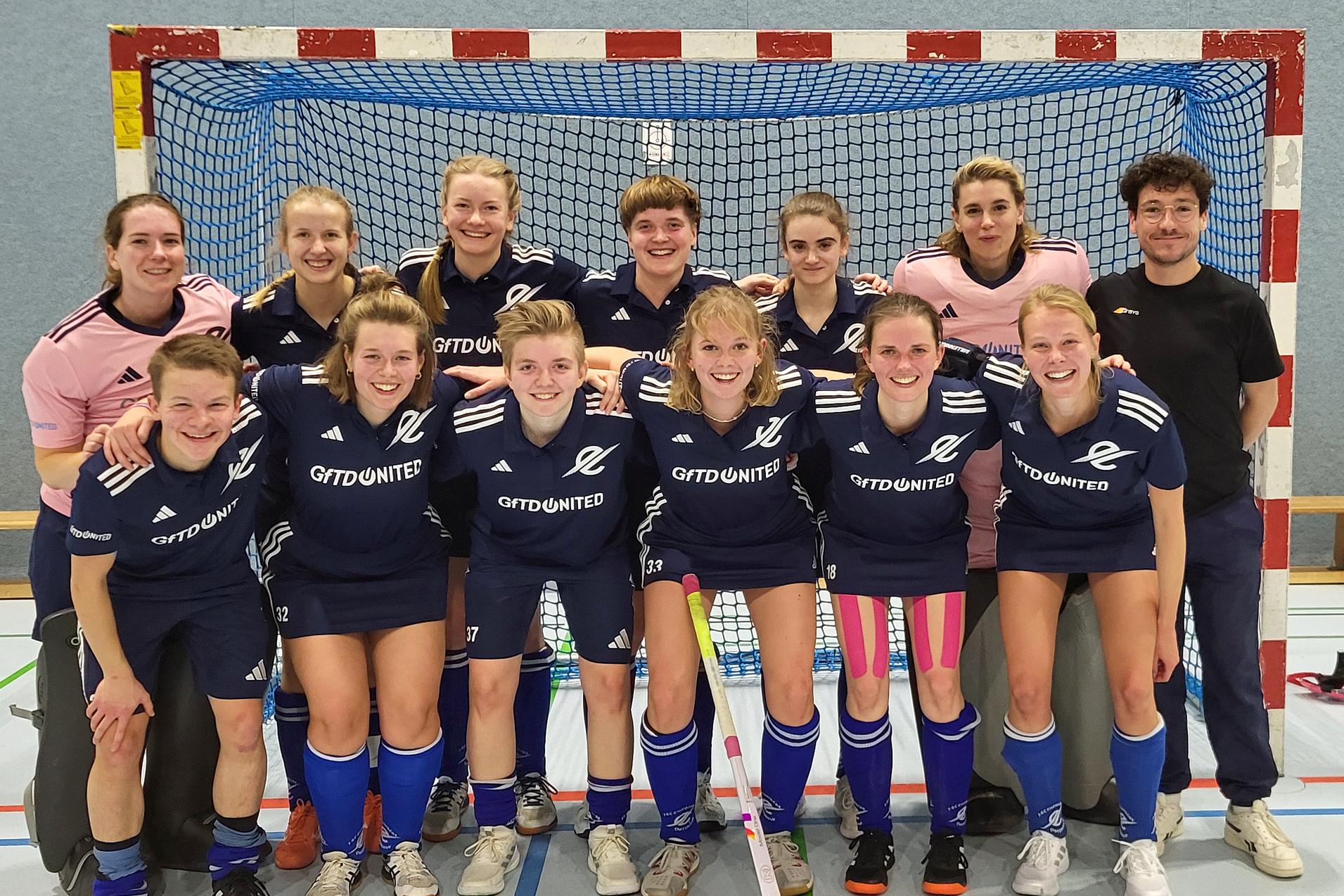 Gruppenfoto einer Frauen-Hockeymannschaft in blauen Trikots vor einem Hockeytor in einer Sporthalle.