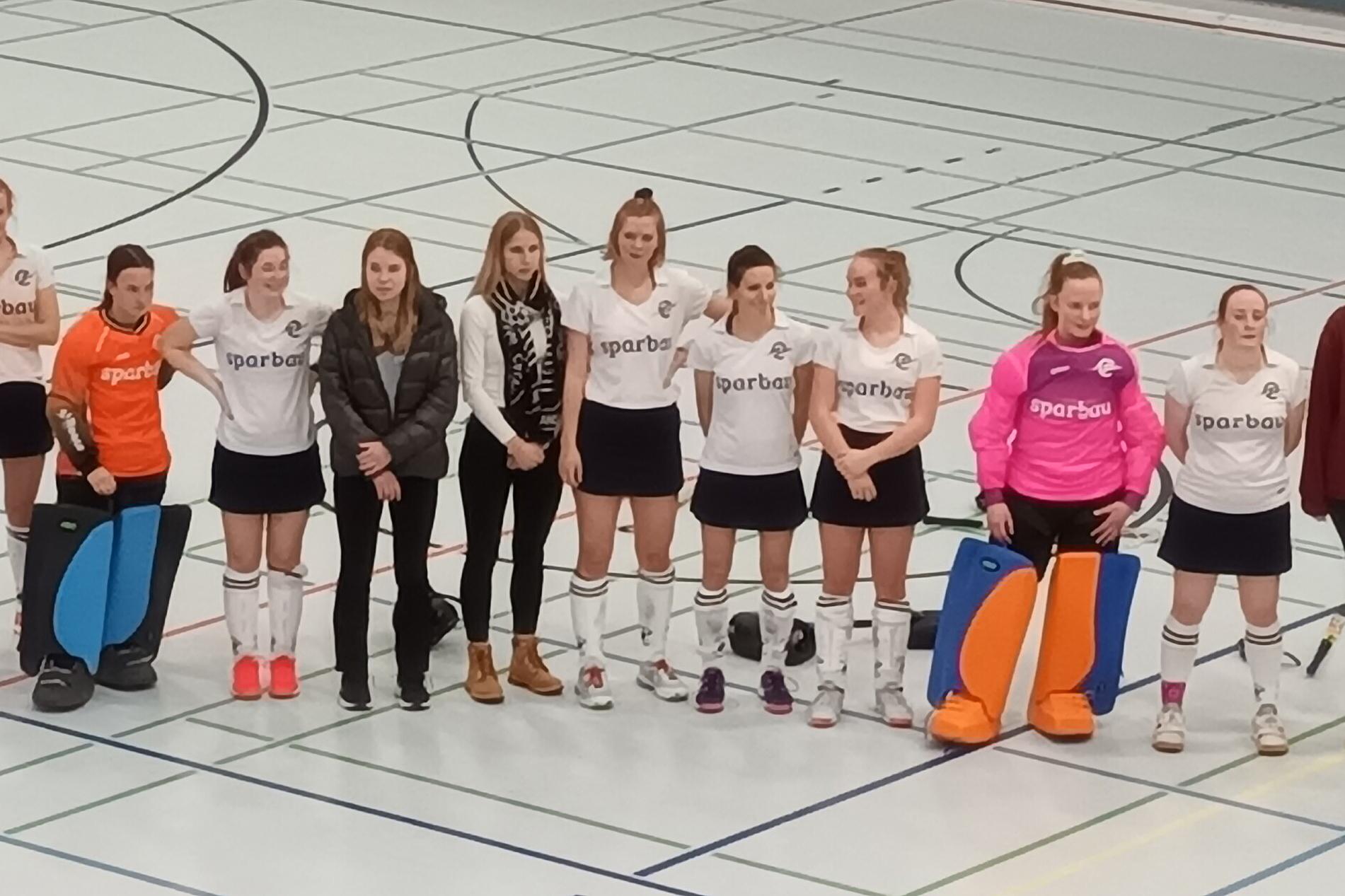 Gruppe von 13 weiblichen Hockeyspielern in Sportkleidung, auf einem Hallenboden mit Linien f&uuml;r Spielfelder.