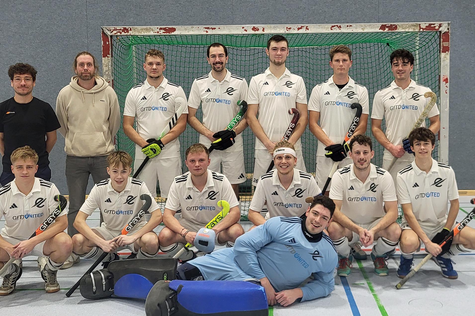 Mannschaftsfoto einer Hockeymannschaft in wei&szlig;en Trikots, aufgenommen vor einem Tor in einer Sporthalle.
