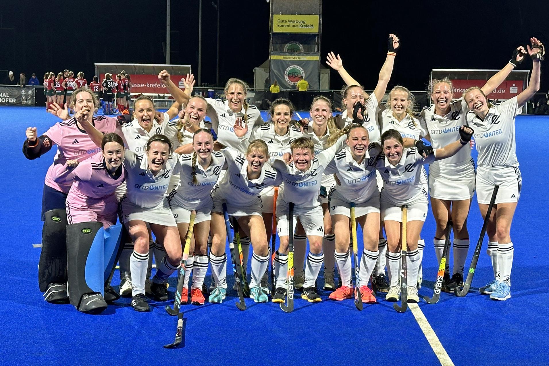 Gruppensiegerin im Feldhockey: 15 Frauen in wei&szlig;en Trikots, jubelnd auf einem blauen Platz, feiern ihren Erfolg.