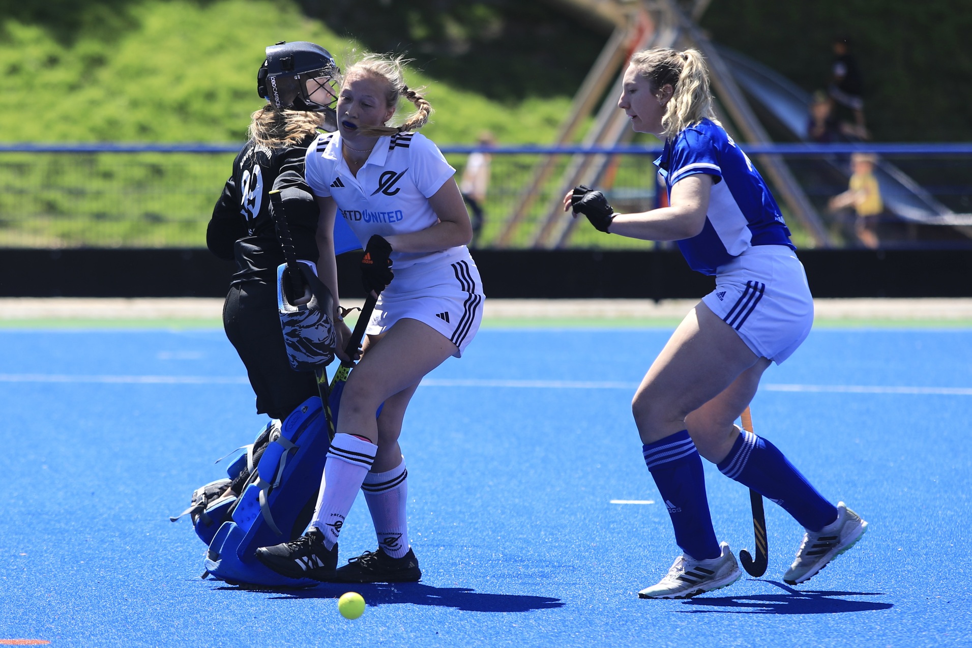 Zwei Hockeyspielerinnen auf einem blauen Kunstrasenfeld, eine dribbelt den Ball, die andere ist im Tor.