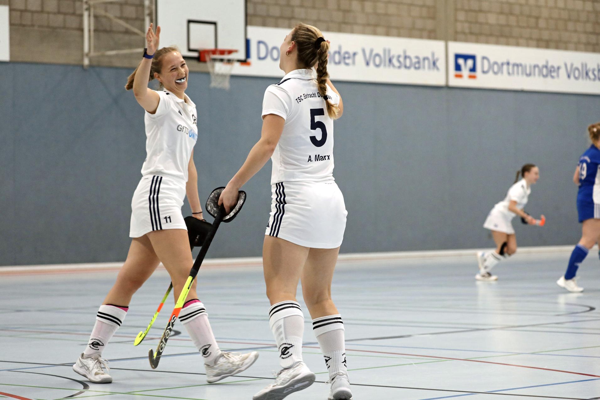 Zwei Hockeyspielerinnen in wei&szlig;en Trikots feiern einen Punkt auf dem Spielfeld in der Halle.