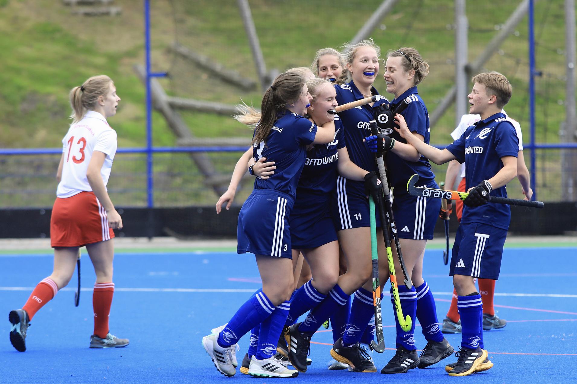 Gruppe junger Hockeyspielerinnen jubelt auf dem Spielfeld, w&auml;hrend eine Spielerin in Wei&szlig; im Hintergrund steht.