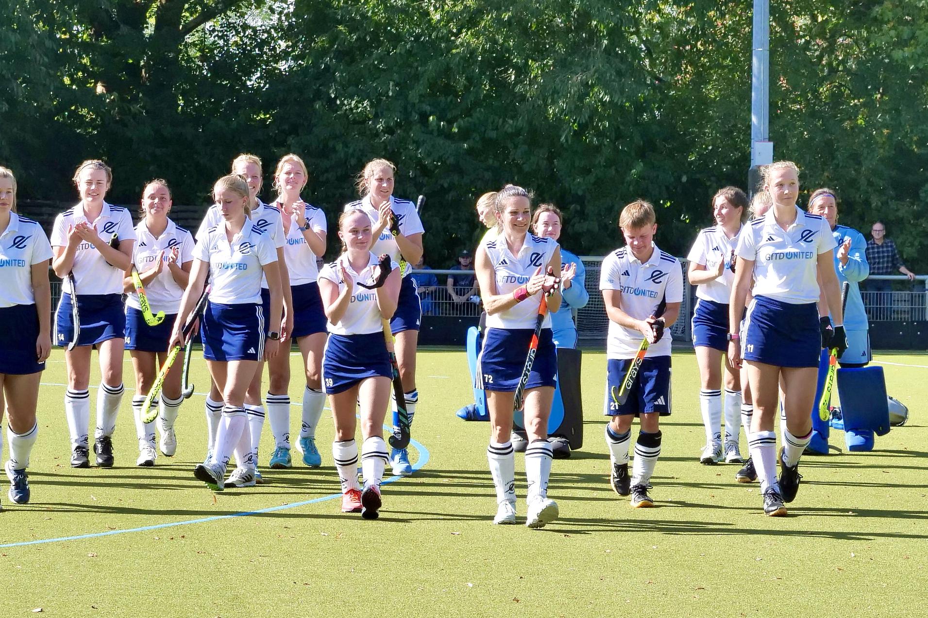 Gruppe von Hockeyspielerinnen in weißen Trikots und blauen Shorts läuft auf einem Kunstrasenfeld.