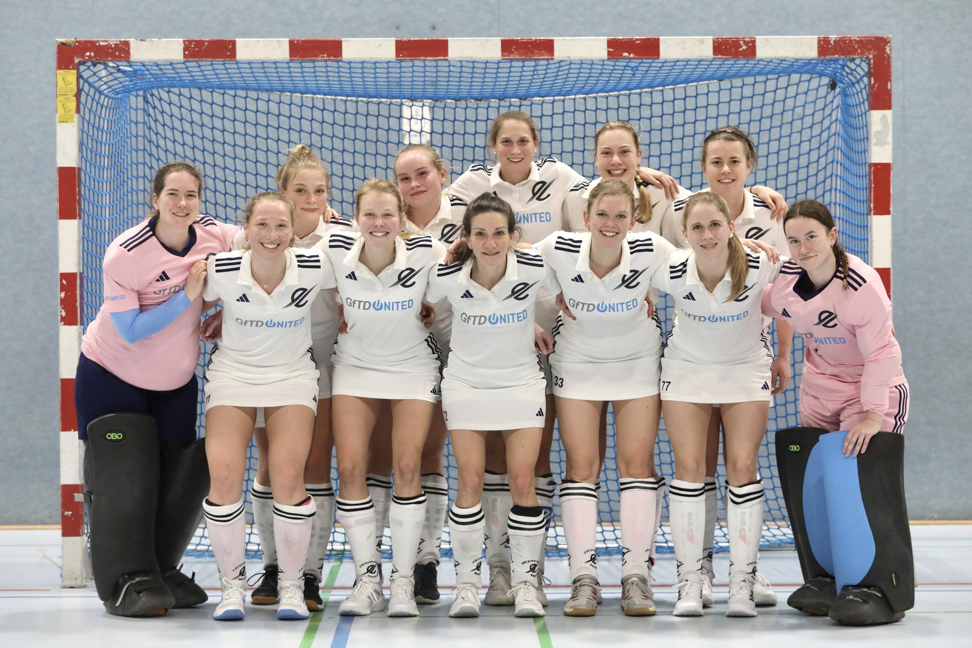 Frauen-Handballmannschaft posiert l&auml;chelnd vor einem Tor in der Halle, einheitlich in wei&szlig;en Trikots gekleidet.