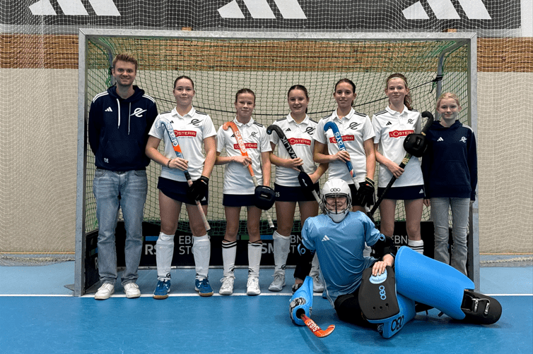 Teamaufstellung von sechs Hockeyspielerinnen und einem Trainer vor einem Tor, eine Spielerin in Torwartausr&uuml;stung sitzt vorne.