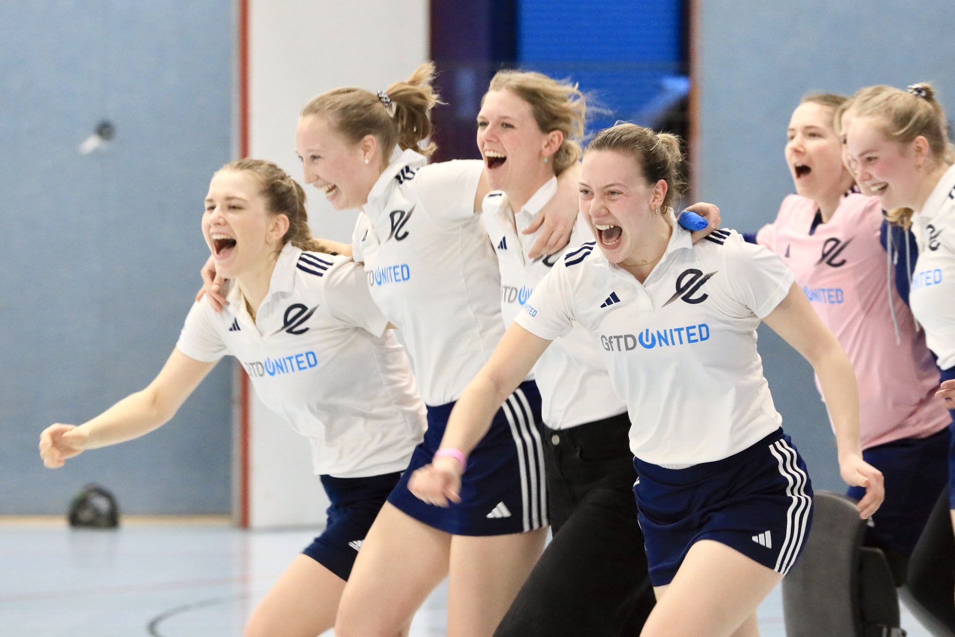 Sechs jubelnde Frauen in Sportkleidung feiern begeistert in einer Halle.