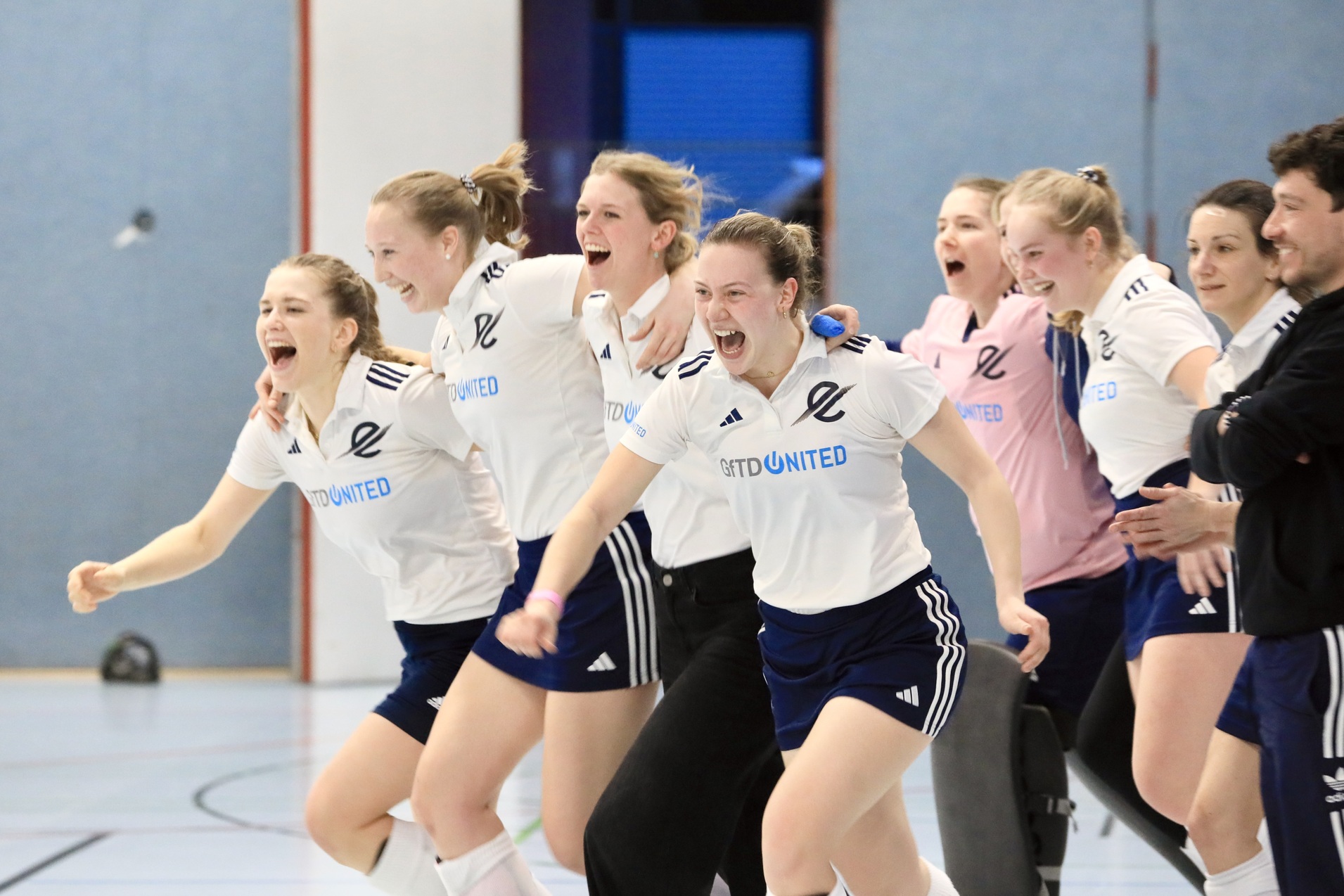 Sechs jubelnde Frauen in Sportkleidung feiern begeistert in einer Halle.