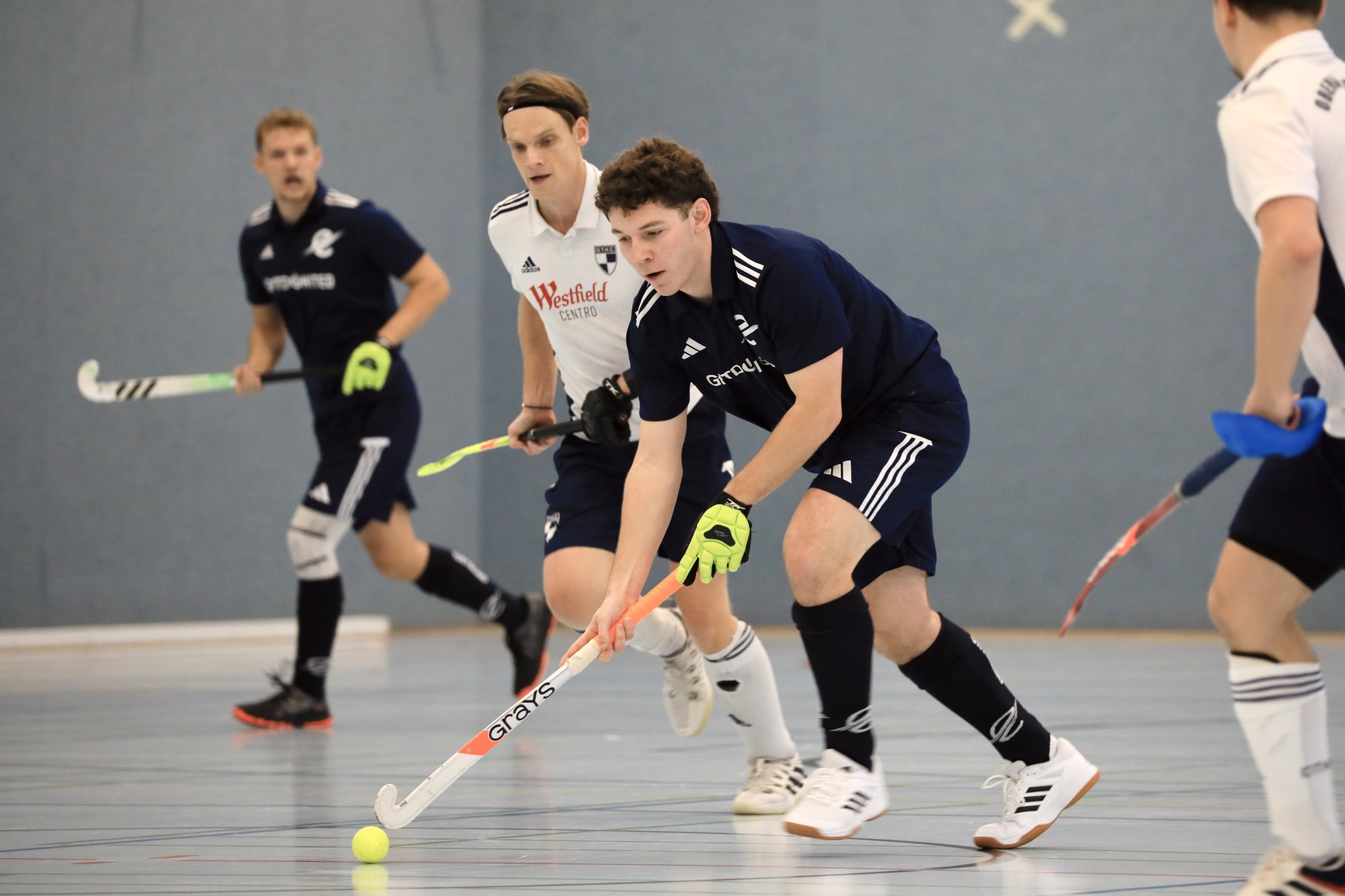 Vier Spieler in Sportkleidung verfolgen einen kleinen Hockeyball auf einem Hallenboden w&auml;hrend eines Spiels.