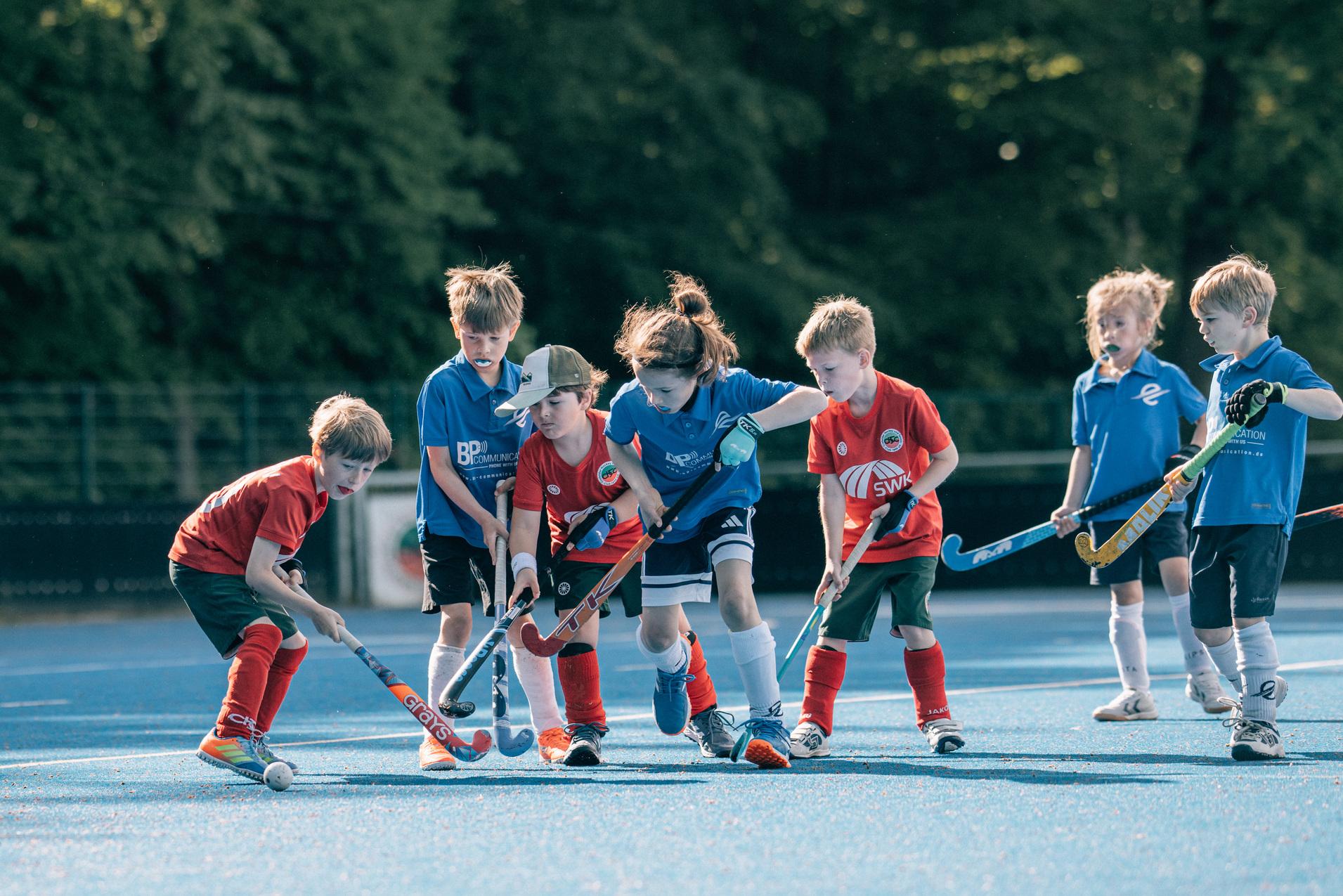 Sechs Kinder mit Hockeyschl&auml;gern spielen auf einem blauen Spielfeld, einige tragen rote, andere blaue Trikots.