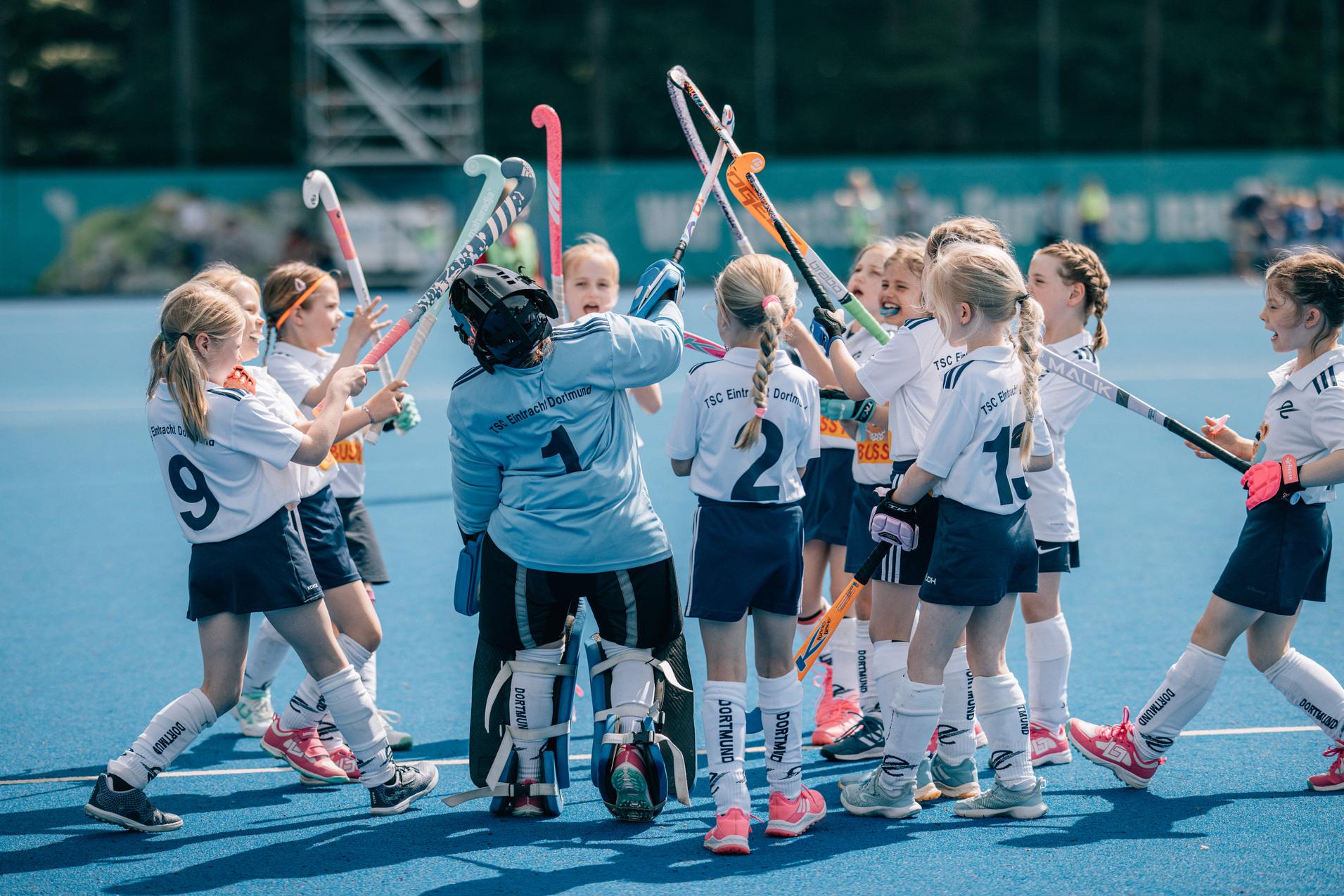 Eine Gruppe junger M&auml;dchen in Sportkleidung umringt eine Torh&uuml;terin auf einem Hockeyfeld. Alle halten Hockeyschl&auml;ger.