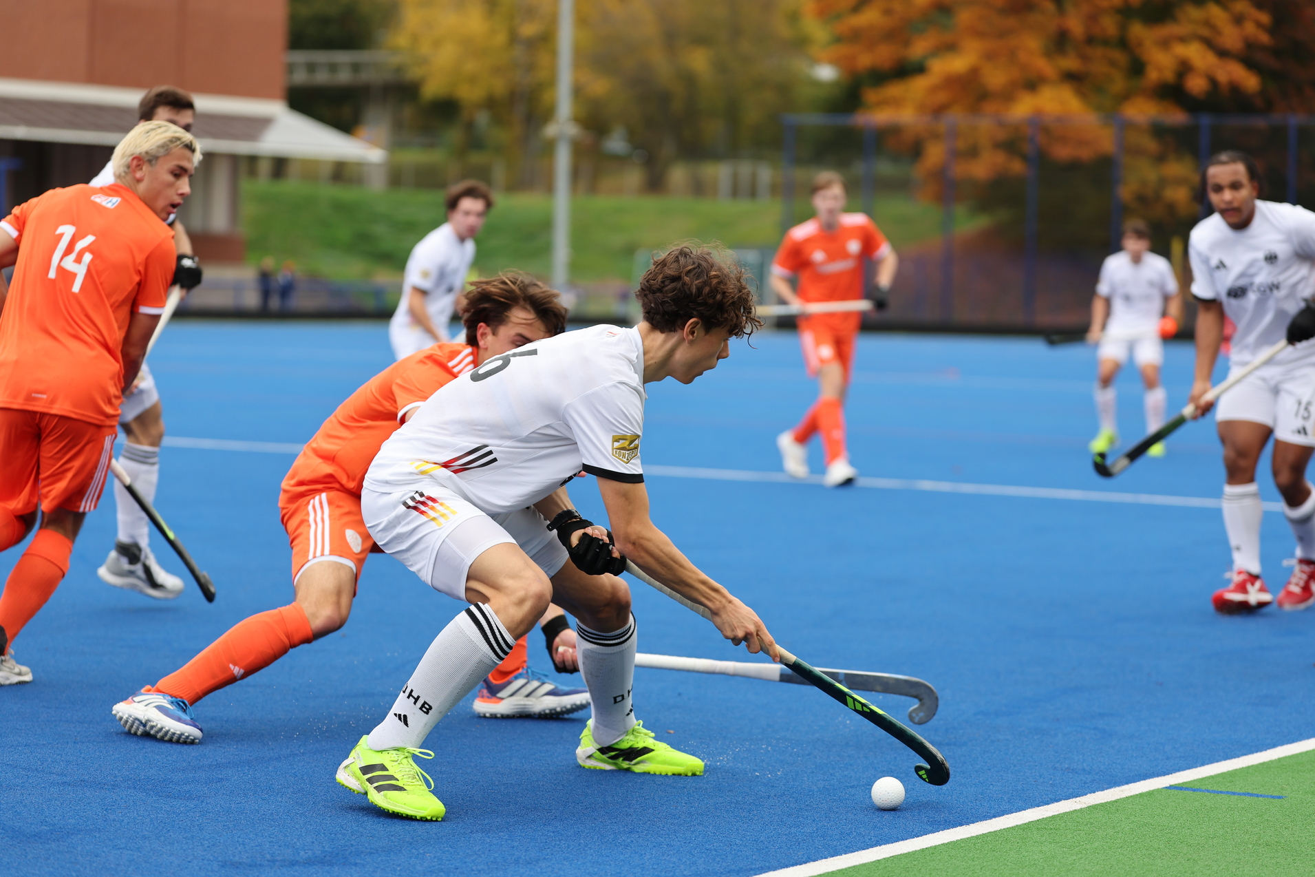 Zwei Hockeyspieler k&auml;mpfen um den Ball auf einem blauen Spielfeld, w&auml;hrend orangefarbene und wei&szlig;e Trikots im Hintergrund sichtbar sind.