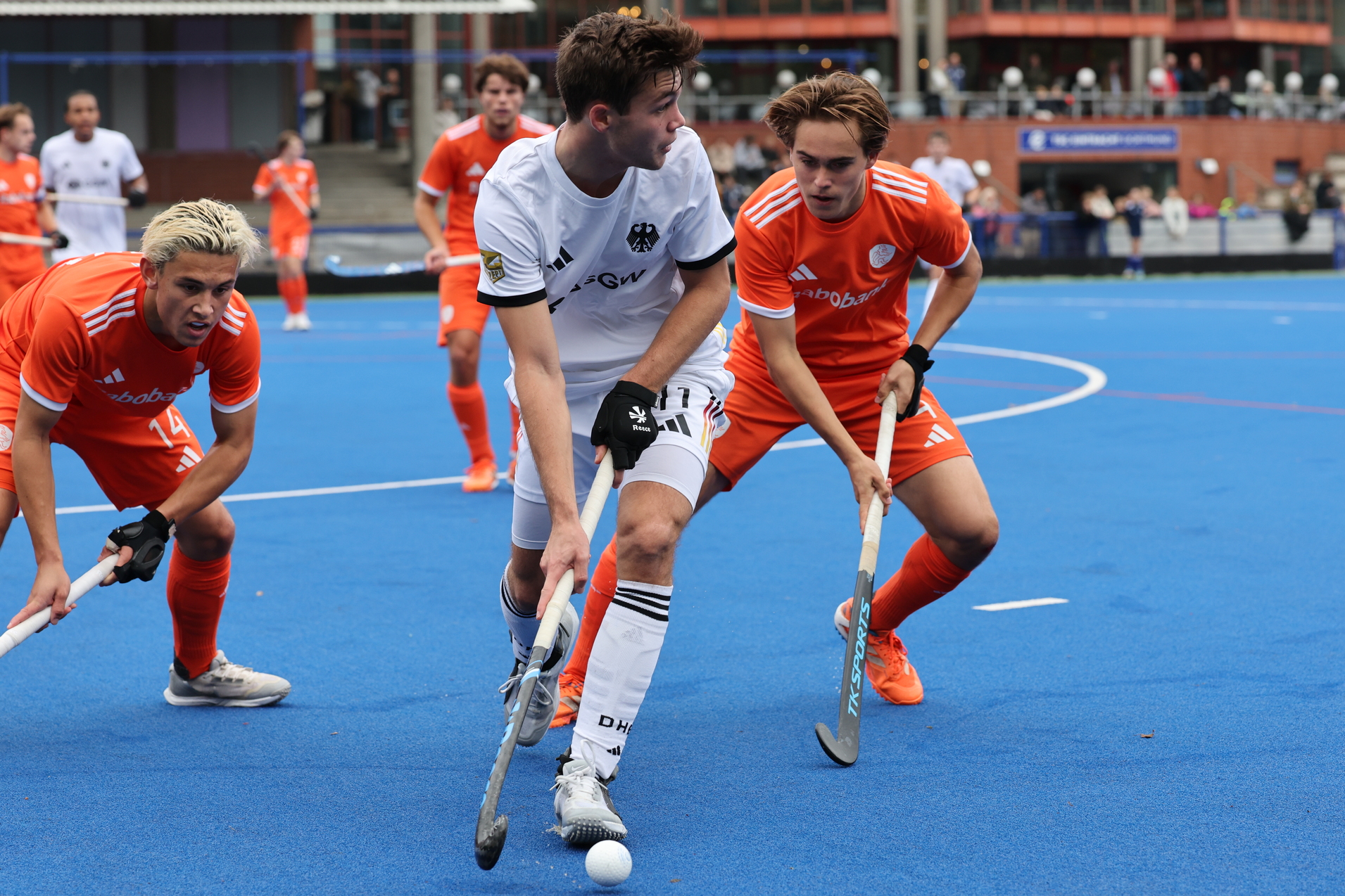 Zwei Spieler in orange und einer in schwarz k&auml;mpfen um den Ball auf einem blauen Hockeyfeld w&auml;hrend eines Spiels.