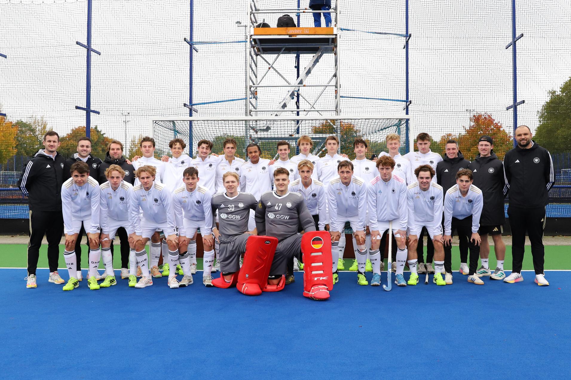 Gruppenzusammenstellung einer Hockeymannschaft in wei&szlig;en Trikots auf einem blauen Feld, Trainer und Spieler l&auml;cheln.