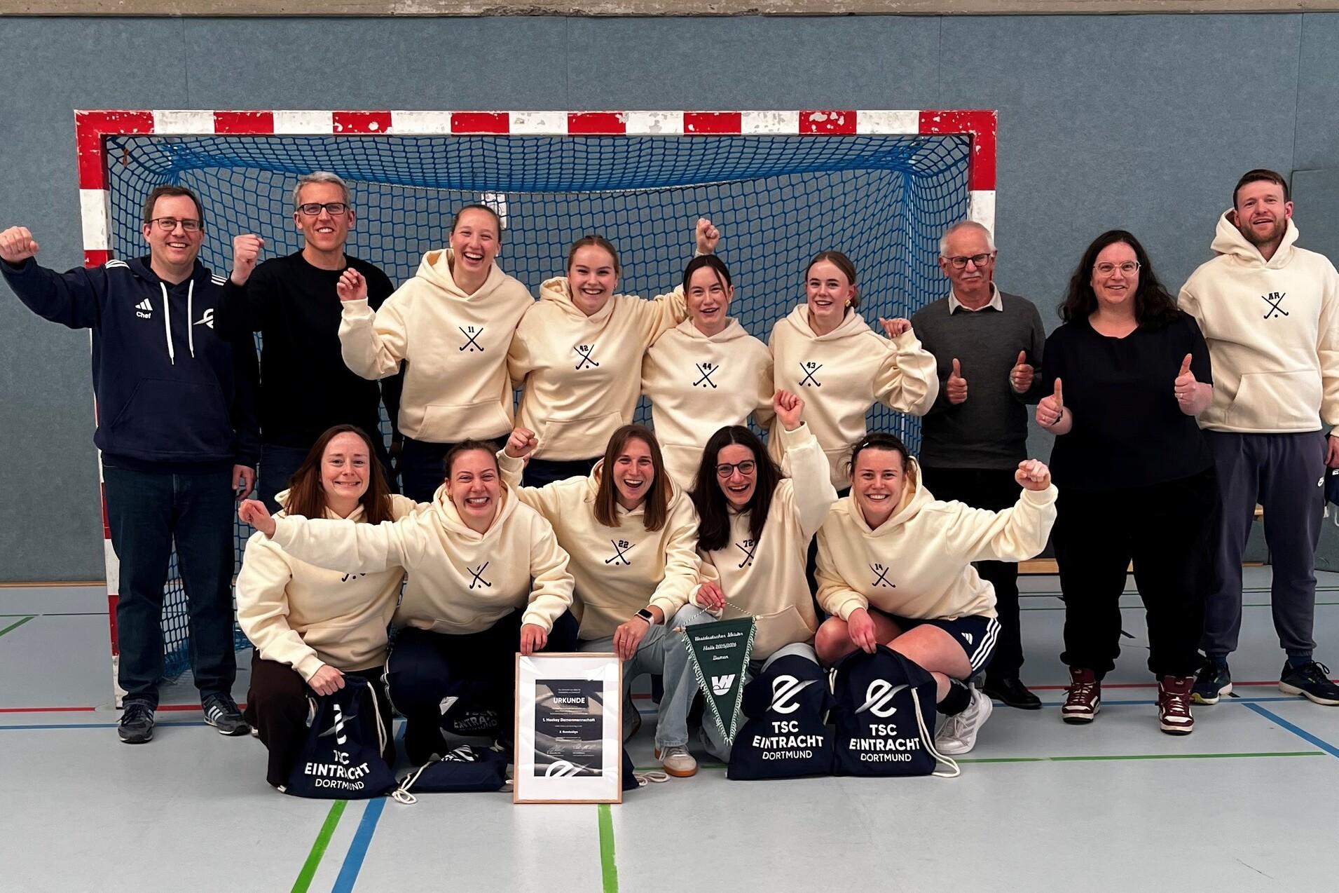 Gruppe von Handballspielern in wei&szlig;en Sweatshirts posiert vor einem Tor, l&auml;chelnd mit Pokalen und Taschen.