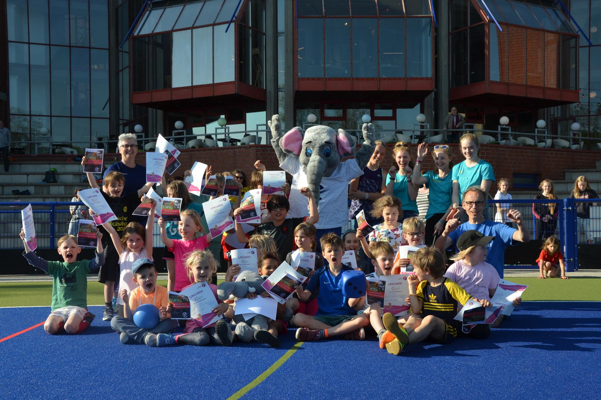 Gruppenfoto von Kindern und Erwachsenen, die stolz Urkunden hochhalten, begleitet von einem Maskottchen auf einem Sportplatz.