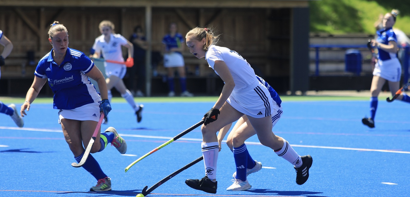 Zwei Hockeyspielerinnen im Wettkampf auf einem blauen Kunstrasenplatz, eine mit Ball, die andere verteidigend.