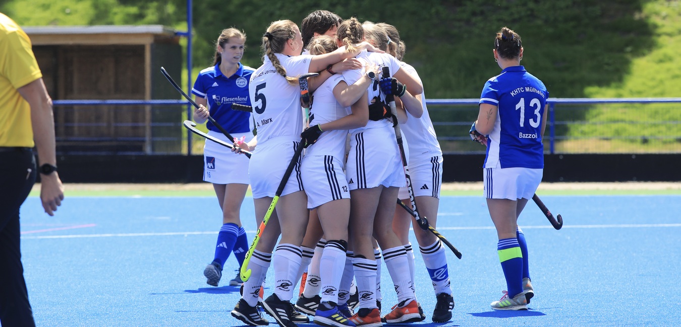 Gruppe von Hockeyspielern in wei&szlig;en Trikots feiert einen Torerfolg auf einem blauen Kunstrasenfeld.