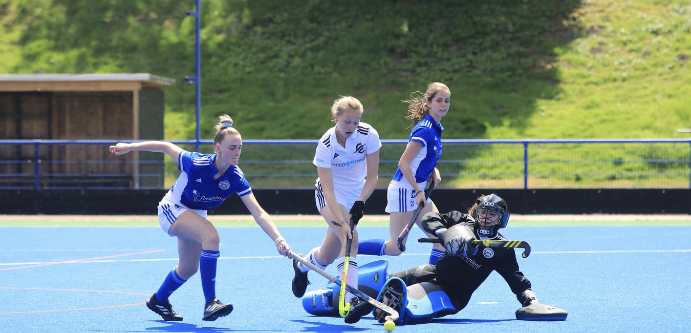 Vier Frauen spielen Hockey auf einem blauen Feld, eine Spielerin schie&szlig;t, w&auml;hrend die Torh&uuml;terin sich abduckt.