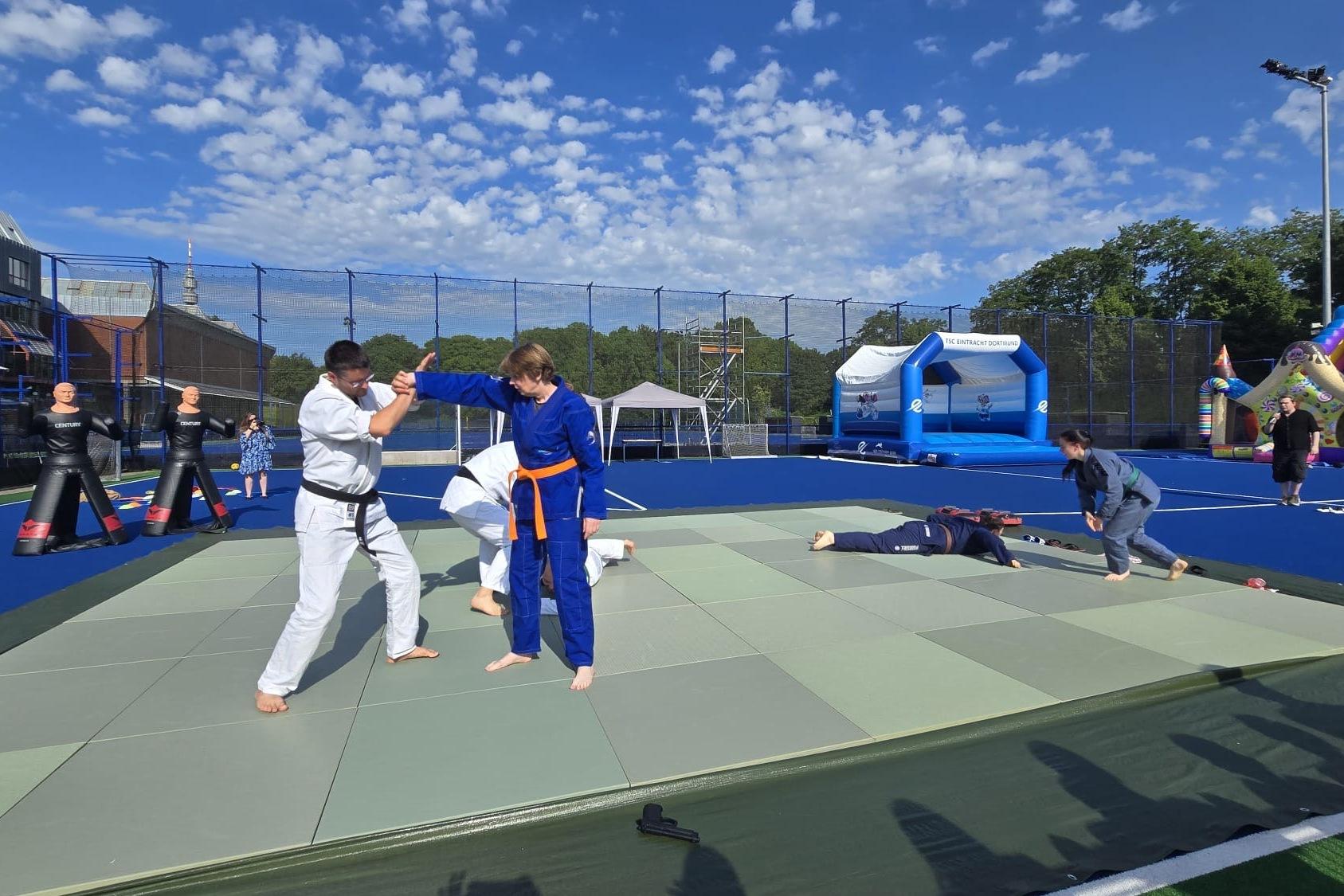 Zwei Judokas in wei&szlig;en und blauen Anz&uuml;gen &uuml;ben auf einer Judomatte, umgeben von spielenden Kindern und einem Spielplatz.