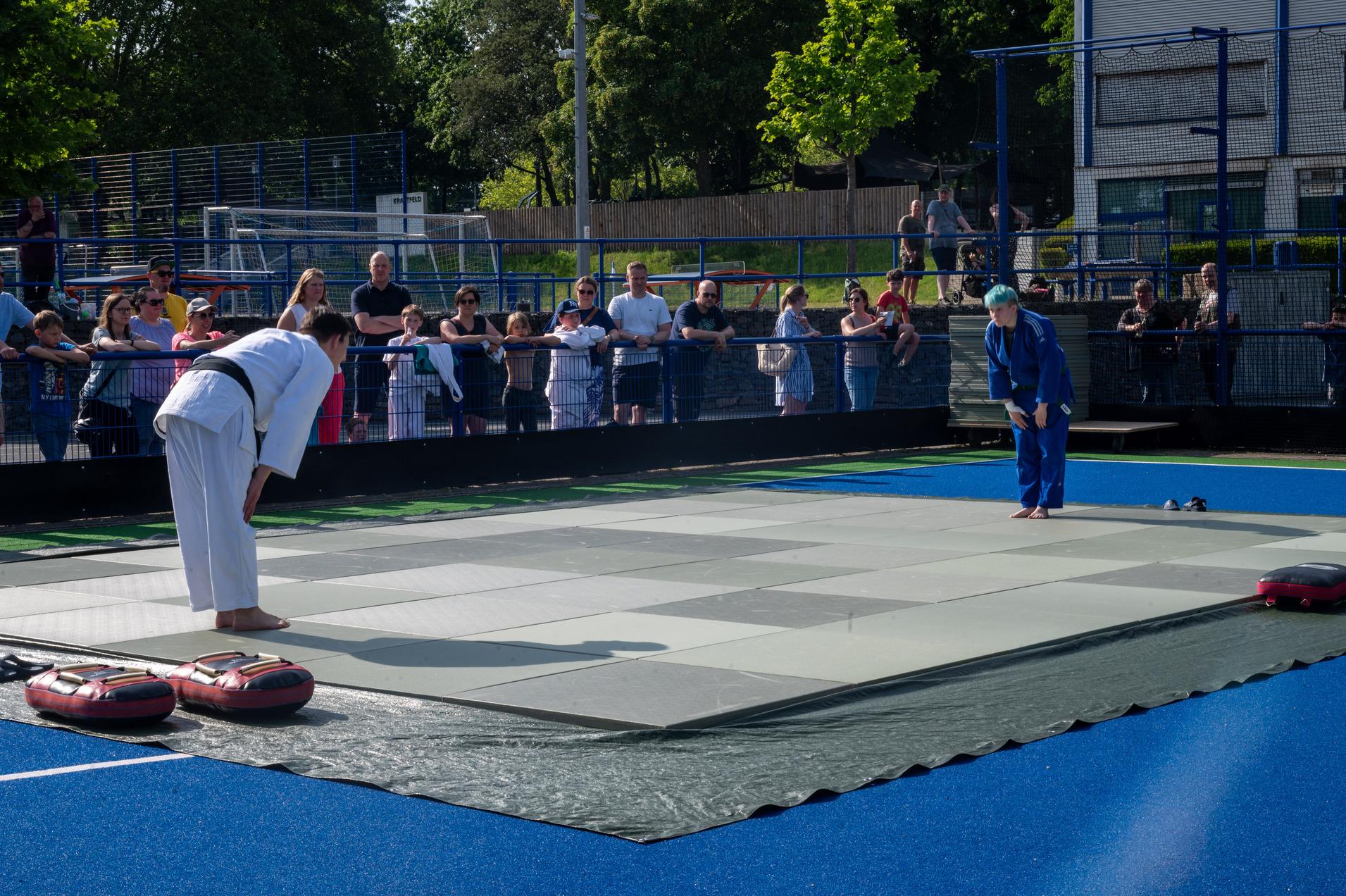 Zwei Judokas in traditioneller Kleidung verbeugen sich auf einer Judomatte, umgeben von Zuschauern.