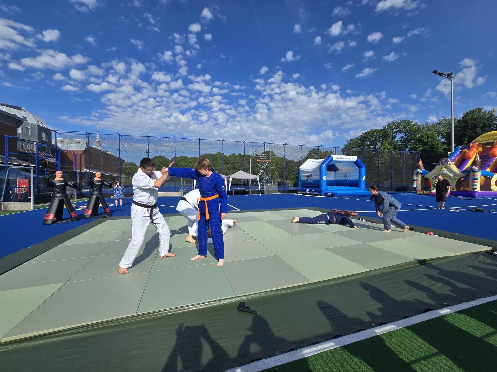 Zwei Judoka im Training auf einer Matte, umgeben von weiteren Sportlern und Spielger&auml;ten im Freien.