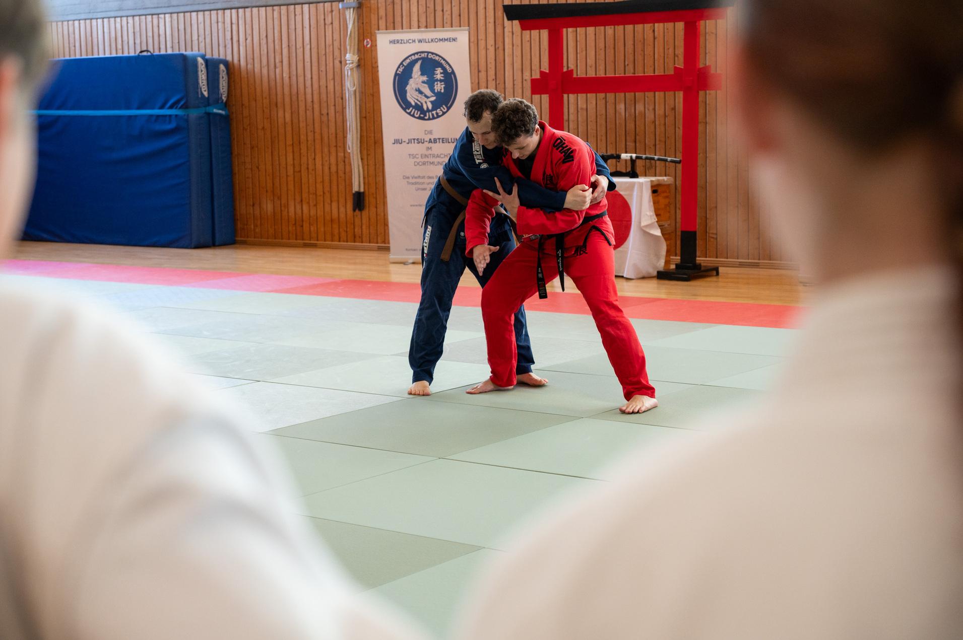 Zwei Judoka, einer in blau und einer in rot, &uuml;ben eine Technik auf einer Judomatte in einem Trainingsraum.