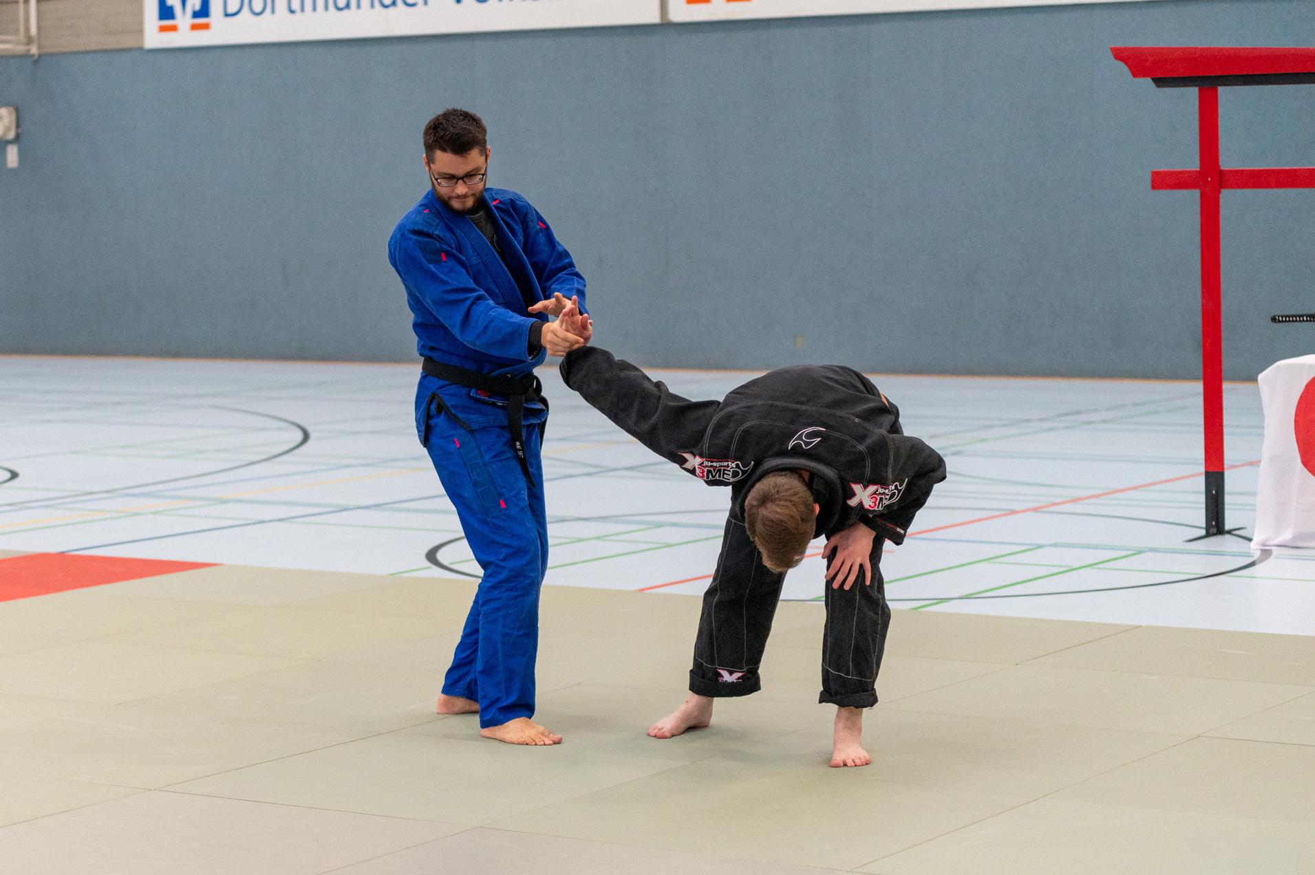 Zwei Kampfsportler &uuml;ben eine Technik auf einer Matte in einer Sporthalle, einer in blauem und der andere in schwarzem Uniform.