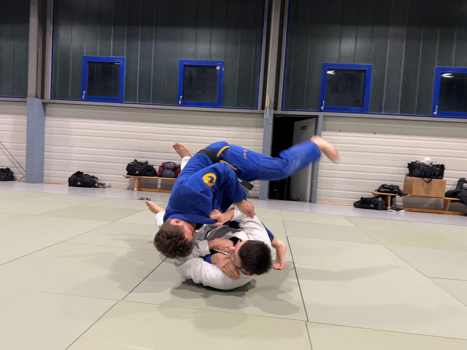 Zwei K&auml;mpfer in BJJ-Uniformen w&auml;hrend eines Wettkampfs auf einer Matte, einer ist im Wurf und der andere am Boden.