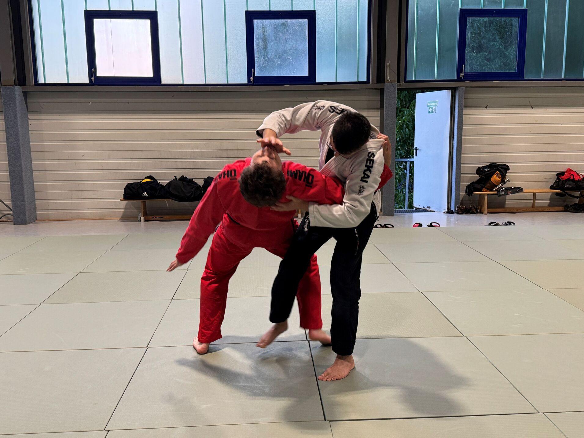 Zwei K&auml;mpfer in Judogis &uuml;ben einen Wurf in einem Trainingsraum mit Mattenbelag.