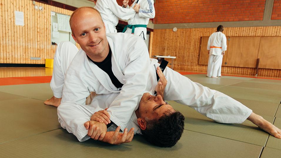 Zwei M&auml;nner in judolike Anz&uuml;gen &uuml;ben in einer Sporthalle einen Halt auf einer Matte.