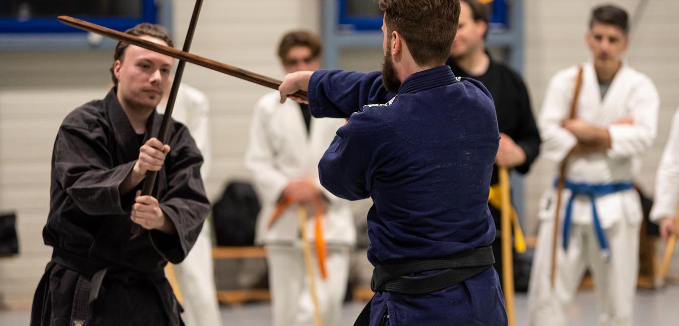 Zwei Kampfsportler in Schwarz und Blau trainieren mit langen St&auml;ben in einem Sportraum, umringt von weiteren Sch&uuml;lern.