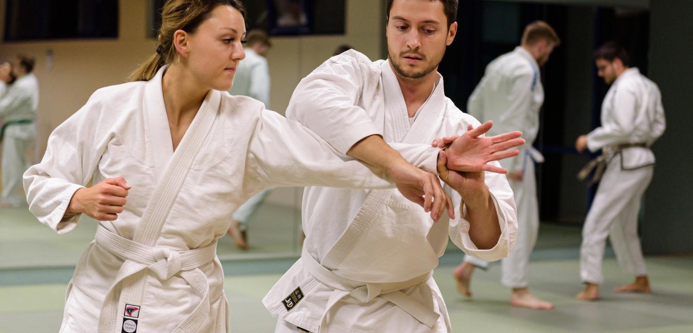 Zwei Judoka in wei&szlig;en Gi-Kits &uuml;ben eine Technik in einem Trainingsraum mit spiegelnden W&auml;nden.