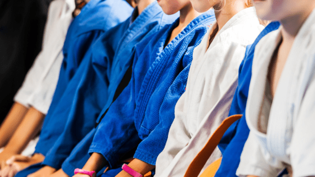Reihe von Kindern im Judoanzug, die im Sitzen auf blauer Matte warten.