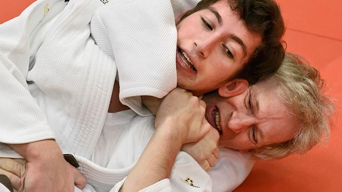 Zwei Judoka im Wettkampf, einer f&uuml;hrt einen W&uuml;rgegriff gegen den anderen auf einer roten Matte aus.