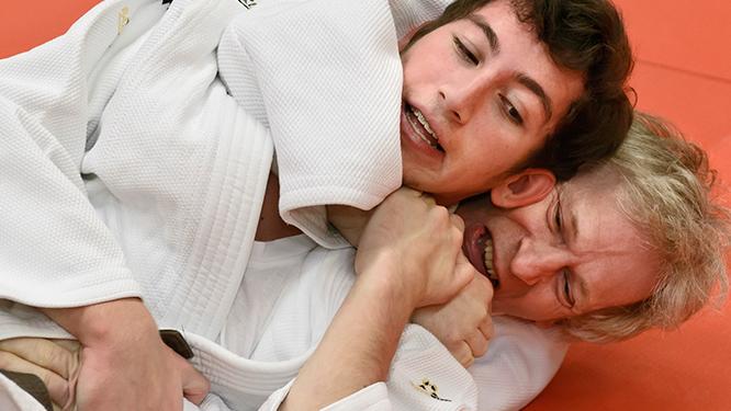 Zwei Judoka im Wettkampf, einer f&uuml;hrt einen W&uuml;rgegriff gegen den anderen auf einer roten Matte aus.
