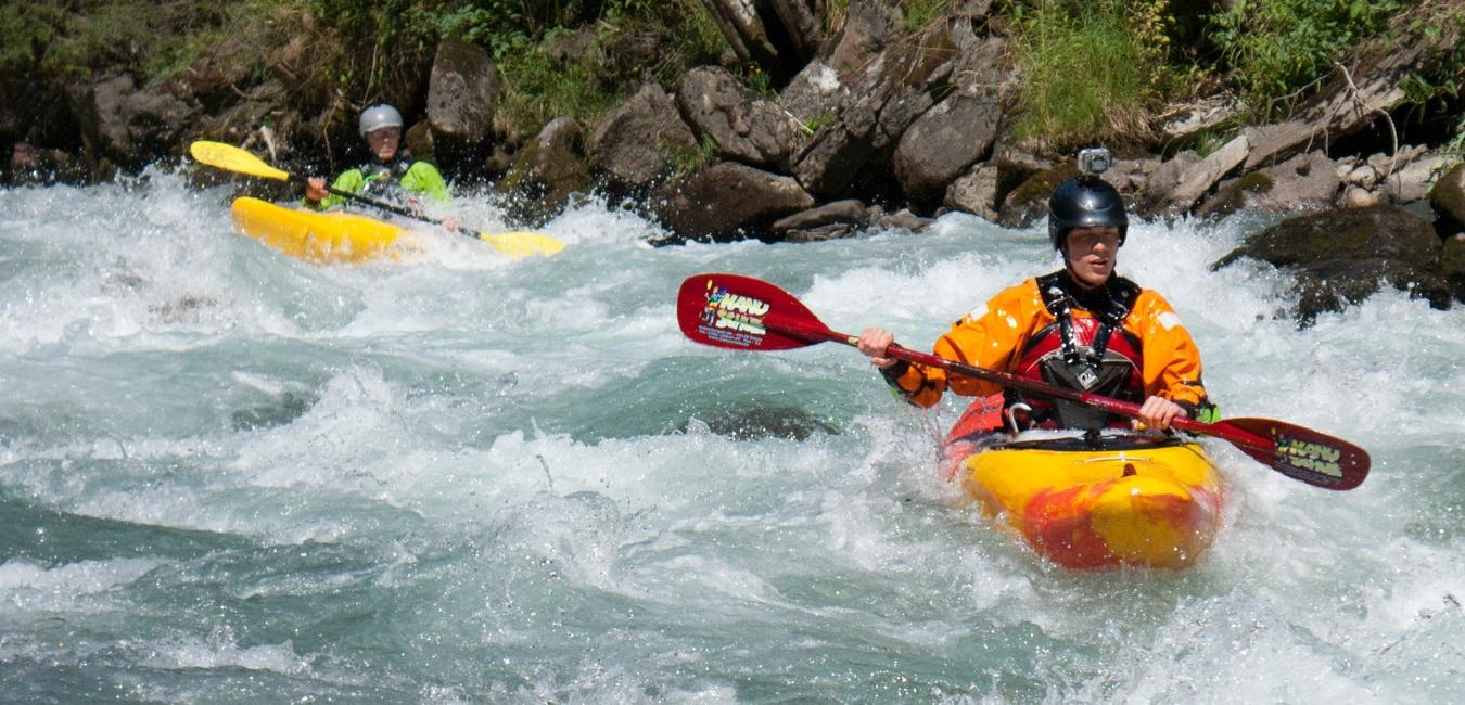 Zwei Kajakfahrer paddeln durch schäumende Wellen in einem Fluss, umgeben von grüner Landschaft.