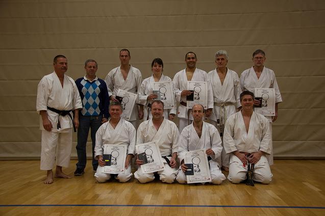 Gruppe von zehn Karateka in Trainingsanz&uuml;gen h&auml;lt Urkunden in einer Sporthalle.